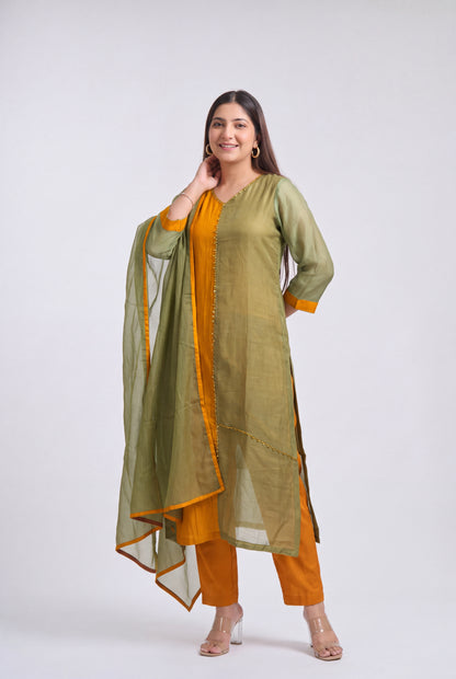 A woman modeling a traditional Indian outfit consisting of an olive green kurta, orange pants, and a matching olive green dupatta with orange border details, set against a plain light gray background.