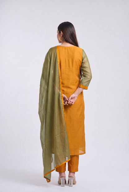 A woman is shown from the back wearing a mustard yellow salwar kameez suit with olive green sleeves and a matching olive green dupatta, set against a plain light gray studio background.