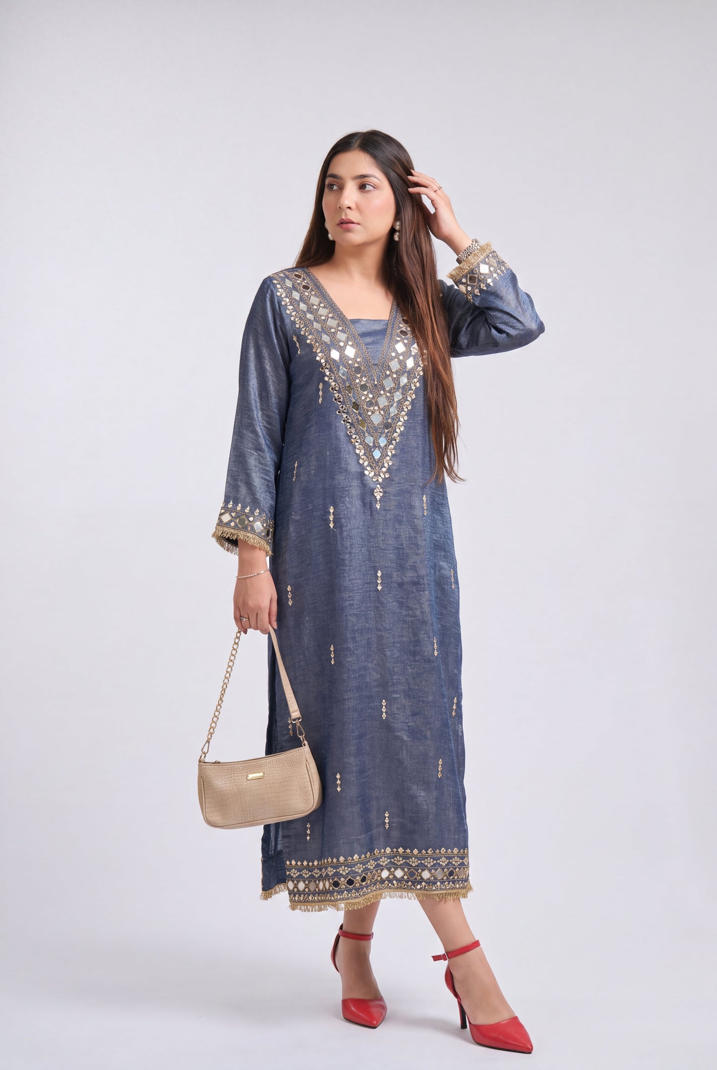 A woman modeling a long-sleeved blue-gray kurta with gold and silver mirror work embroidery around the neckline, cuffs, and hem, paired with red high heels and a beige handbag, against a light gray studio background.