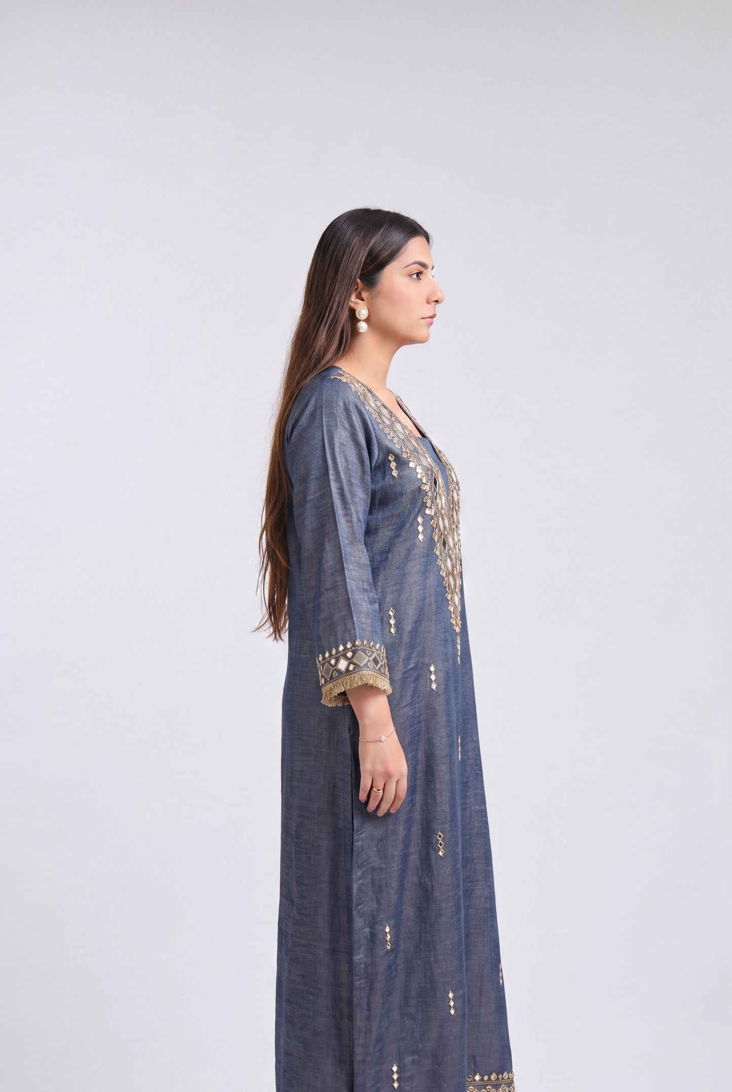 A woman stands in profile wearing a long blue-gray kurta with gold embroidery and mirror work on the neckline, cuffs, and hem, paired with white heeled sandals against a light gray studio background.
