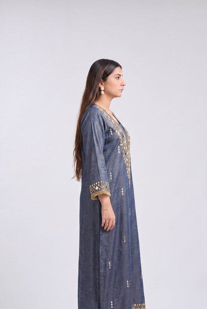 A woman stands in profile wearing a long blue-gray kurta with gold embroidery and mirror work on the neckline, cuffs, and hem, paired with white heeled sandals against a light gray studio background.