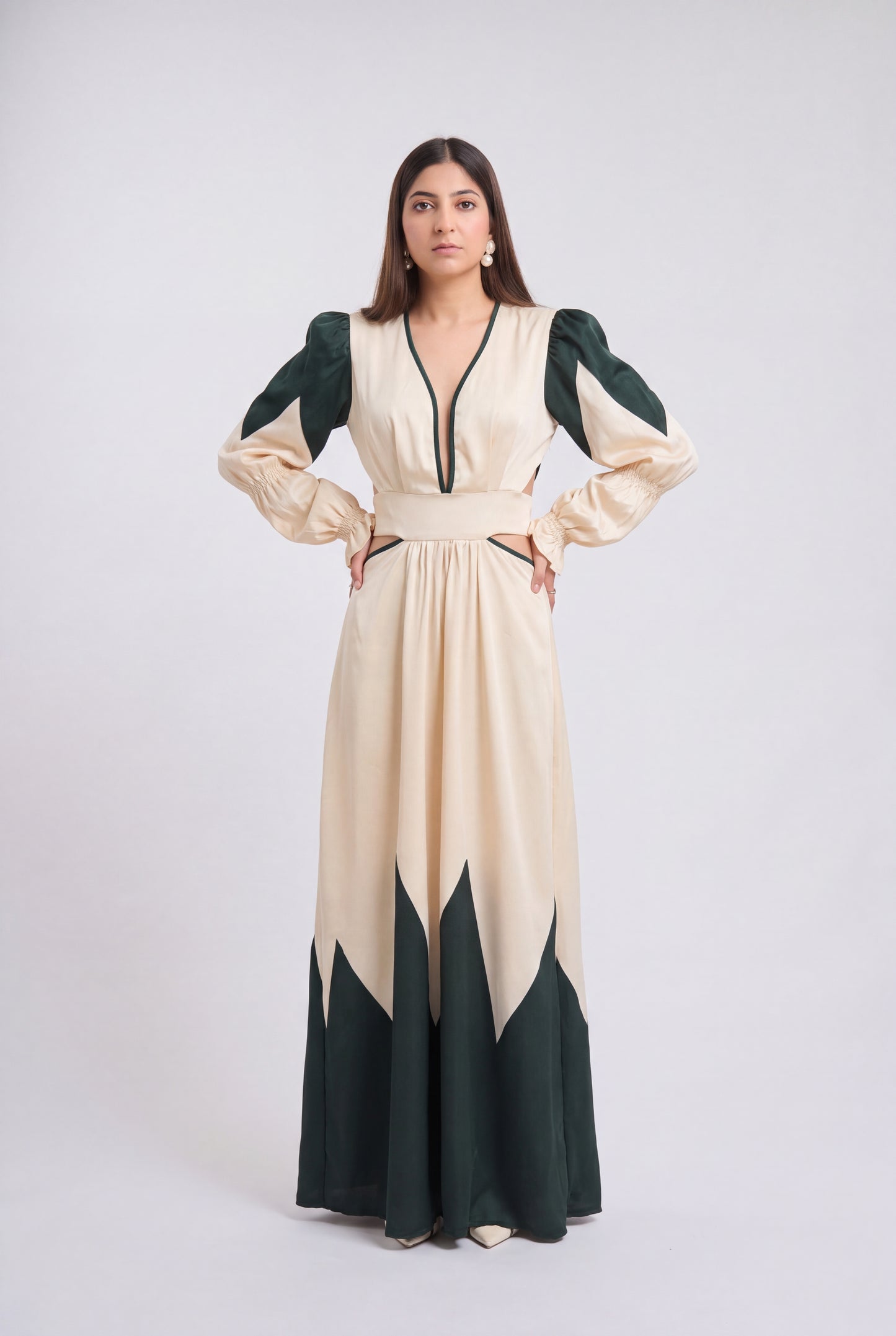 A woman modeling a long cream maxi dress with dark green color-blocked sleeves, neckline trim, and hem details, standing against a light gray studio background.