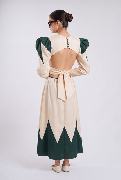 A woman shown from the back wearing a long cream dress with dark green contrast panels, puffed long sleeves, and a tied open back, photographed against a light gray studio background.