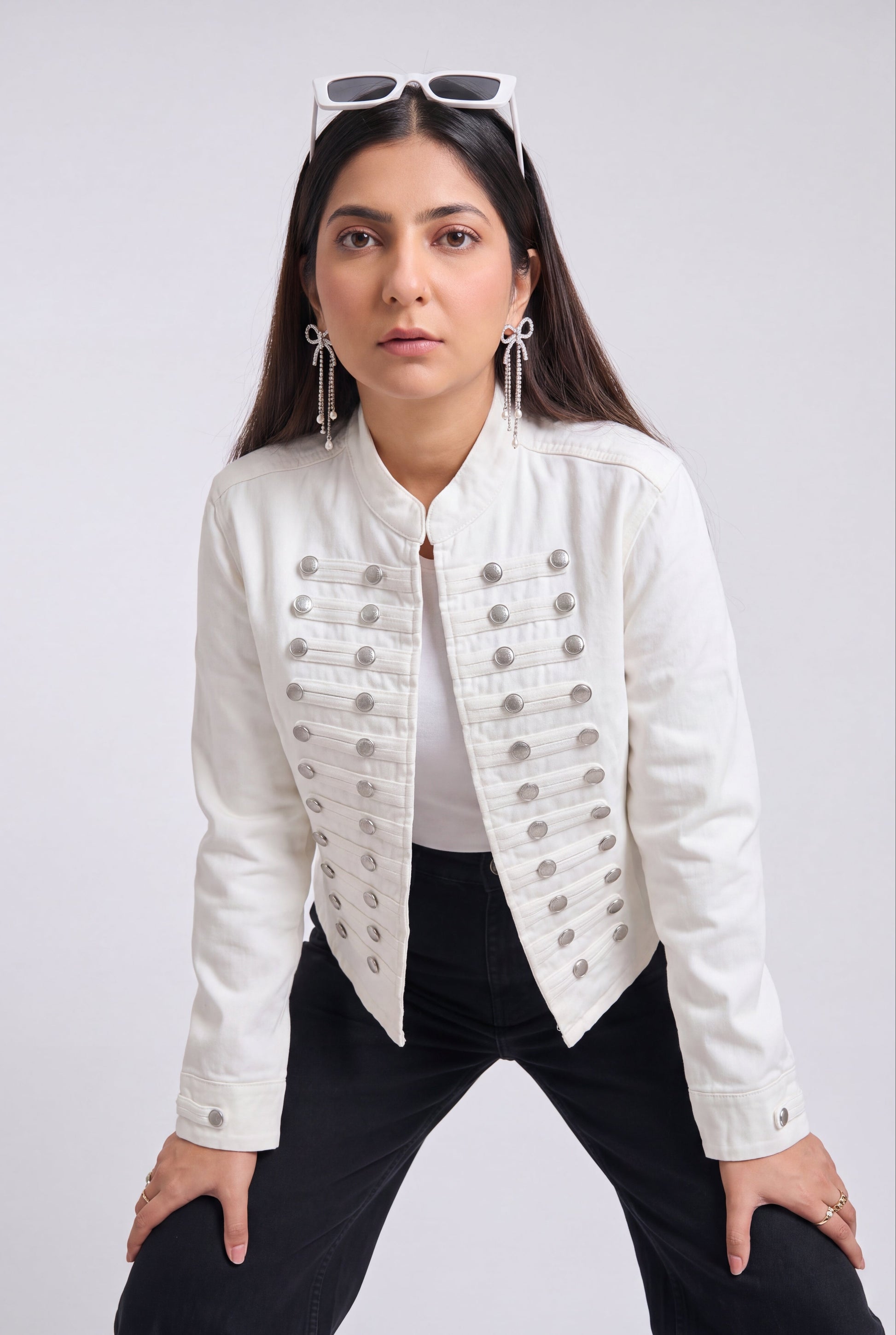 A woman modeling a white cropped jacket with silver military-style buttons, paired with black pants and white sunglasses on her head, against a light gray studio background.