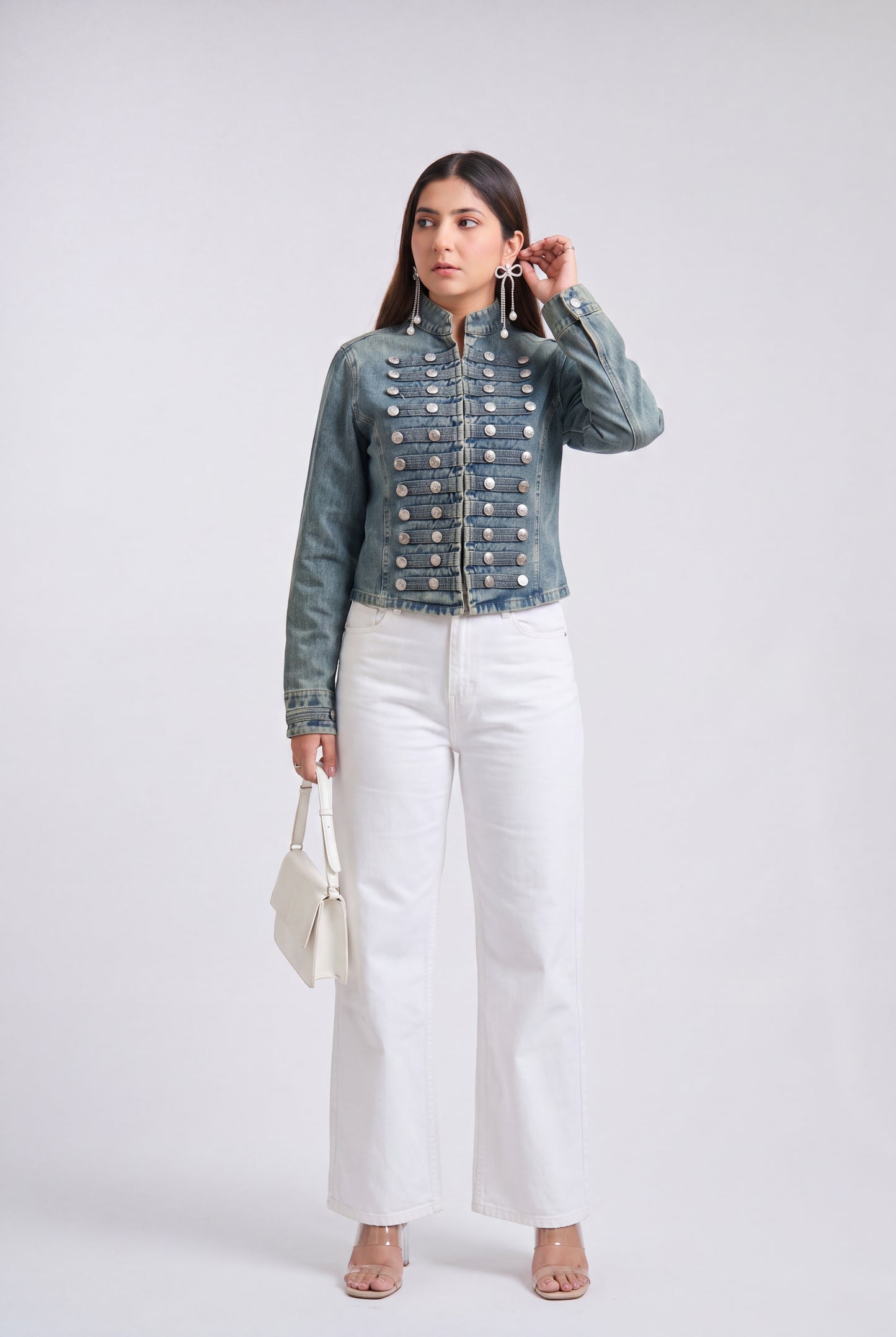 A woman modeling an denim blue military-style jacket with silver buttons, paired with black wide-leg pants and clear high-heeled sandals, against a light gray studio background.