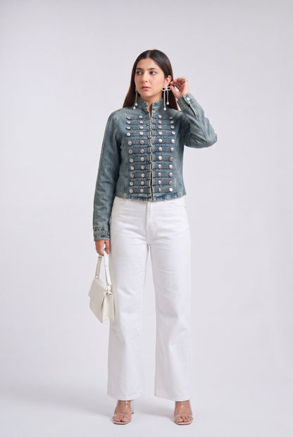 A woman modeling an denim blue military-style jacket with silver buttons, paired with black wide-leg pants and clear high-heeled sandals, against a light gray studio background.