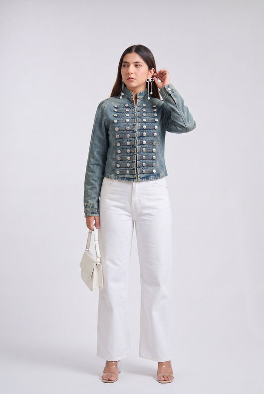 A woman modeling an denim blue military-style jacket with silver buttons, paired with black wide-leg pants and clear high-heeled sandals, against a light gray studio background.