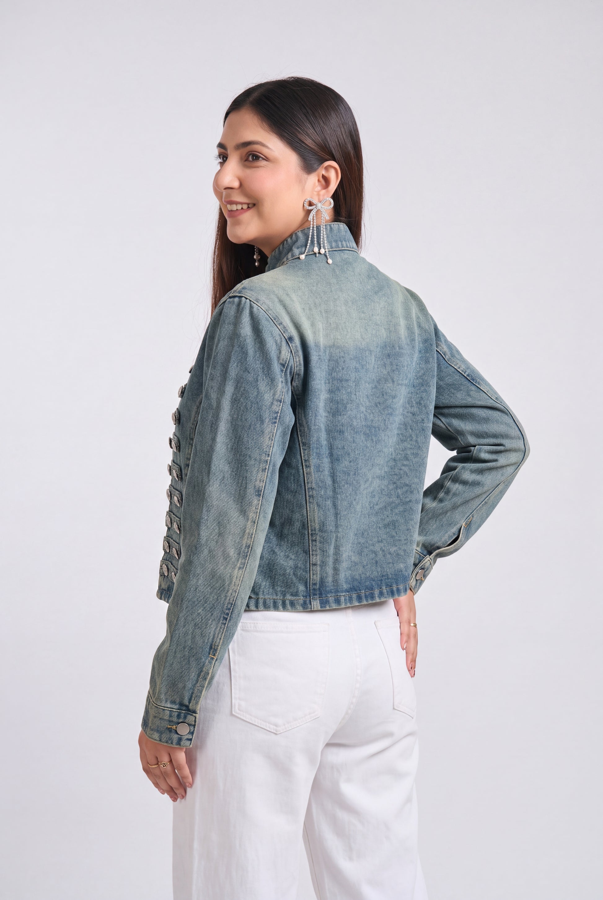 A woman wearing a cropped, light-wash blue denim jacket with decorative lace-up detailing on the back, paired with white pants, photographed against a light gray studio background.