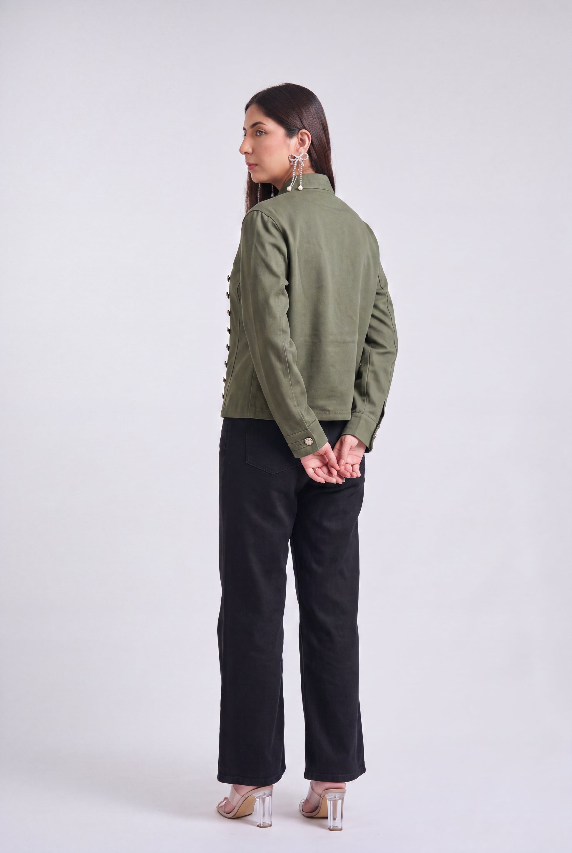 A woman modeling an olive green military-style jacket with silver buttons, paired with black wide-leg pants and clear high-heeled sandals, against a light gray studio background.