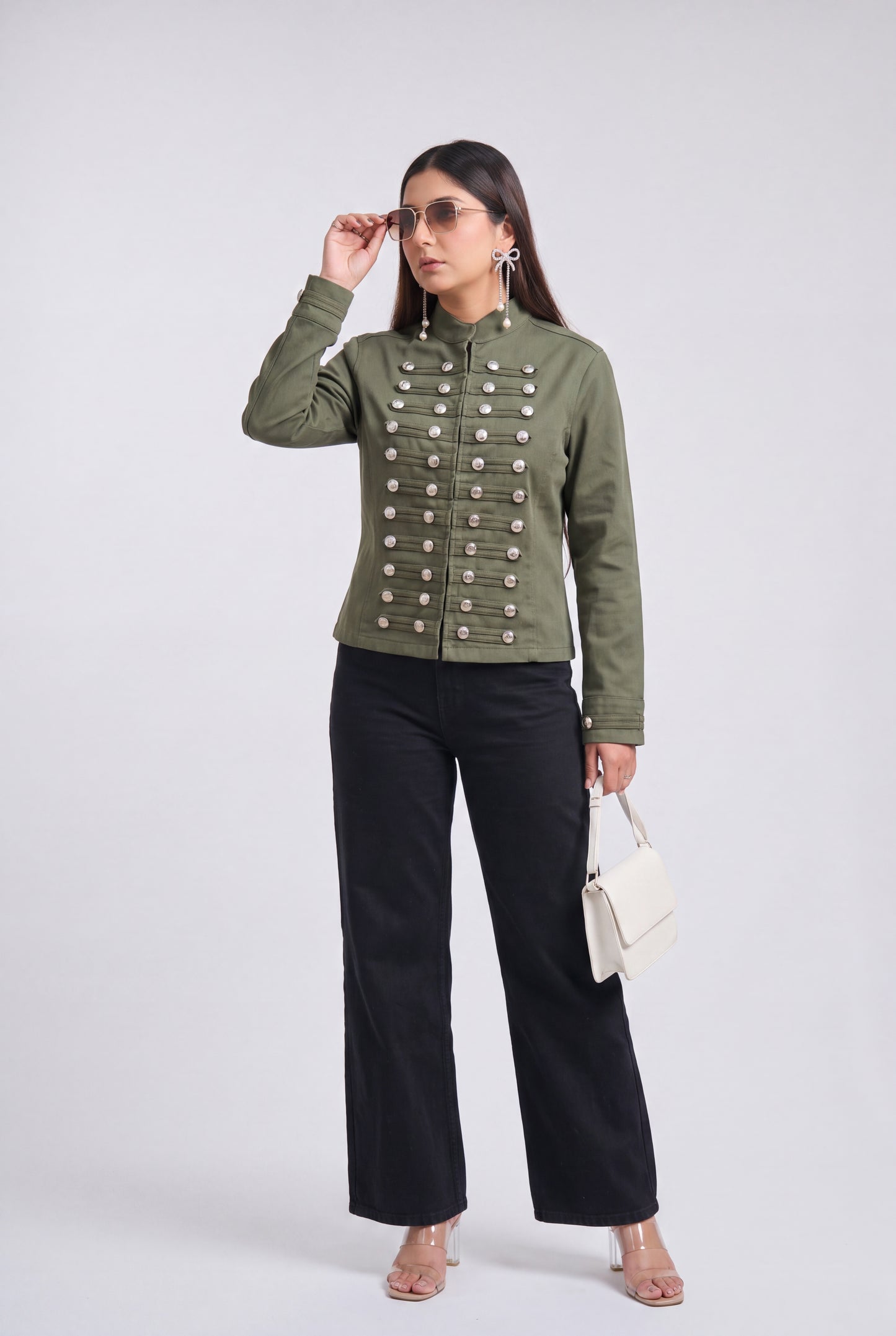 A woman modeling an olive green military-style jacket with silver buttons, paired with black wide-leg pants and clear high-heeled sandals, against a light gray studio background.