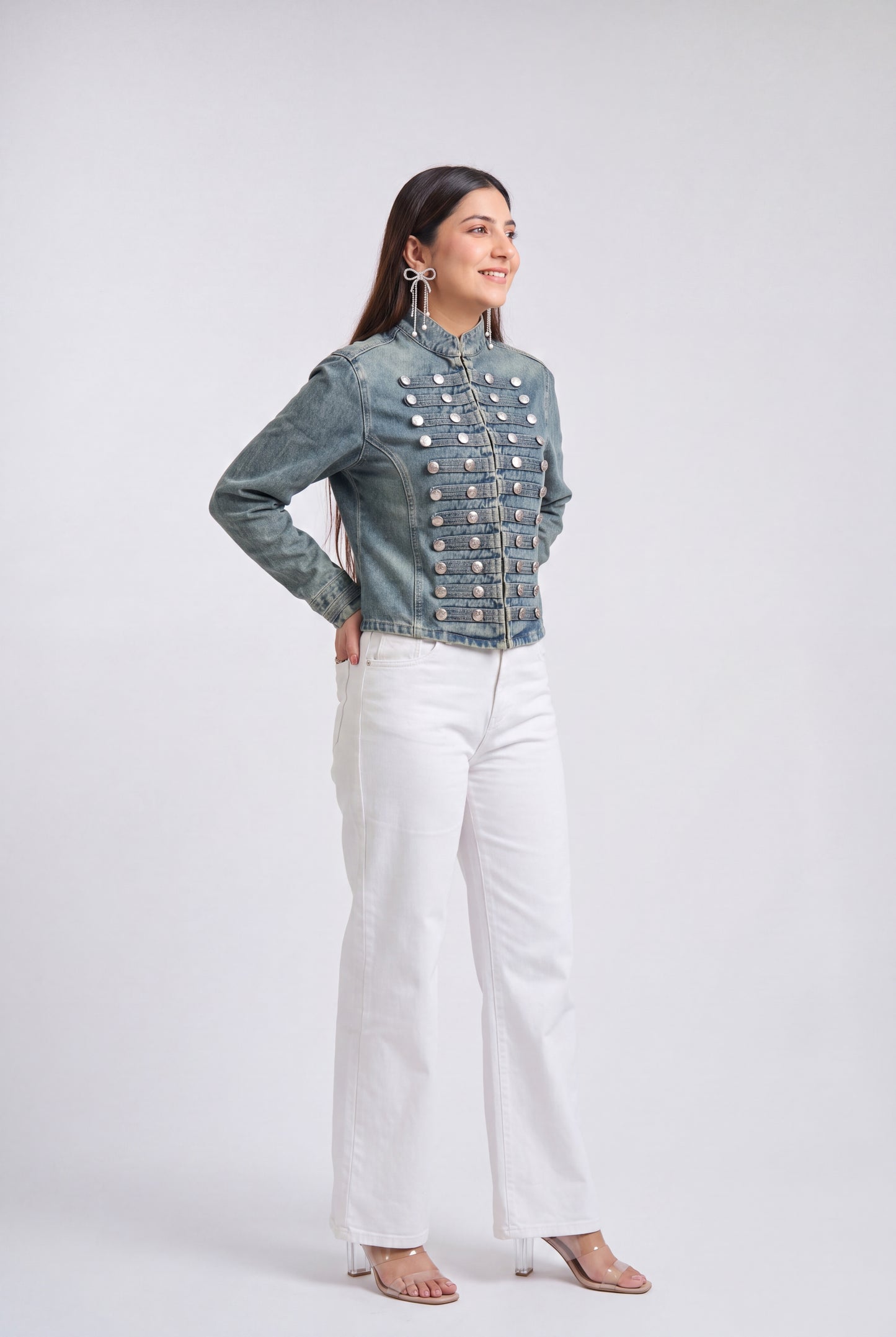 A woman modeling a blue denim jacket with ornate silver military-style snap and button detailing across the chest, paired with white wide-leg pants and clear high-heeled sandals, against a light gray studio background.