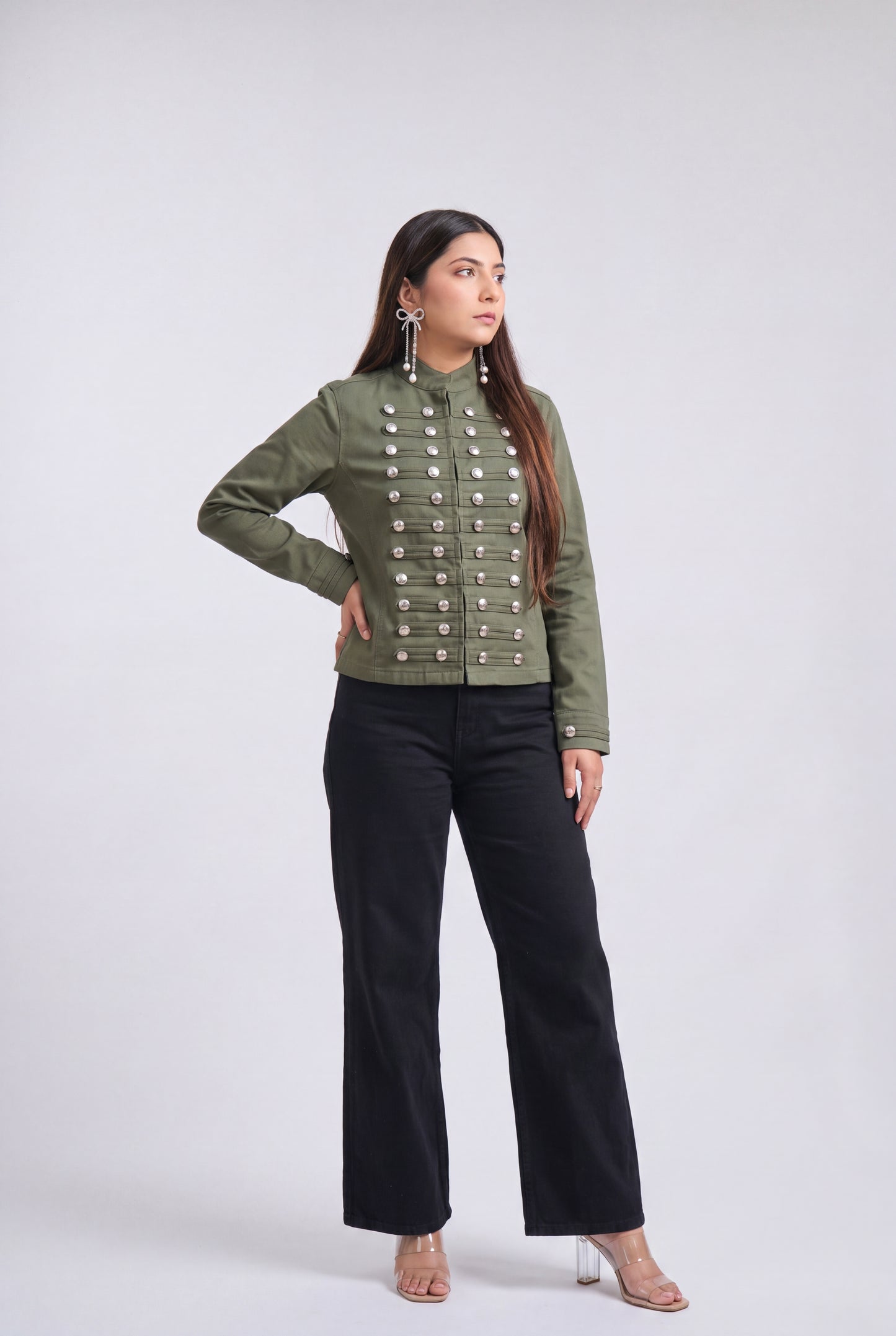 A woman modeling an olive green military-style jacket with silver buttons, paired with black wide-leg pants and clear high-heeled sandals, against a light gray studio background.