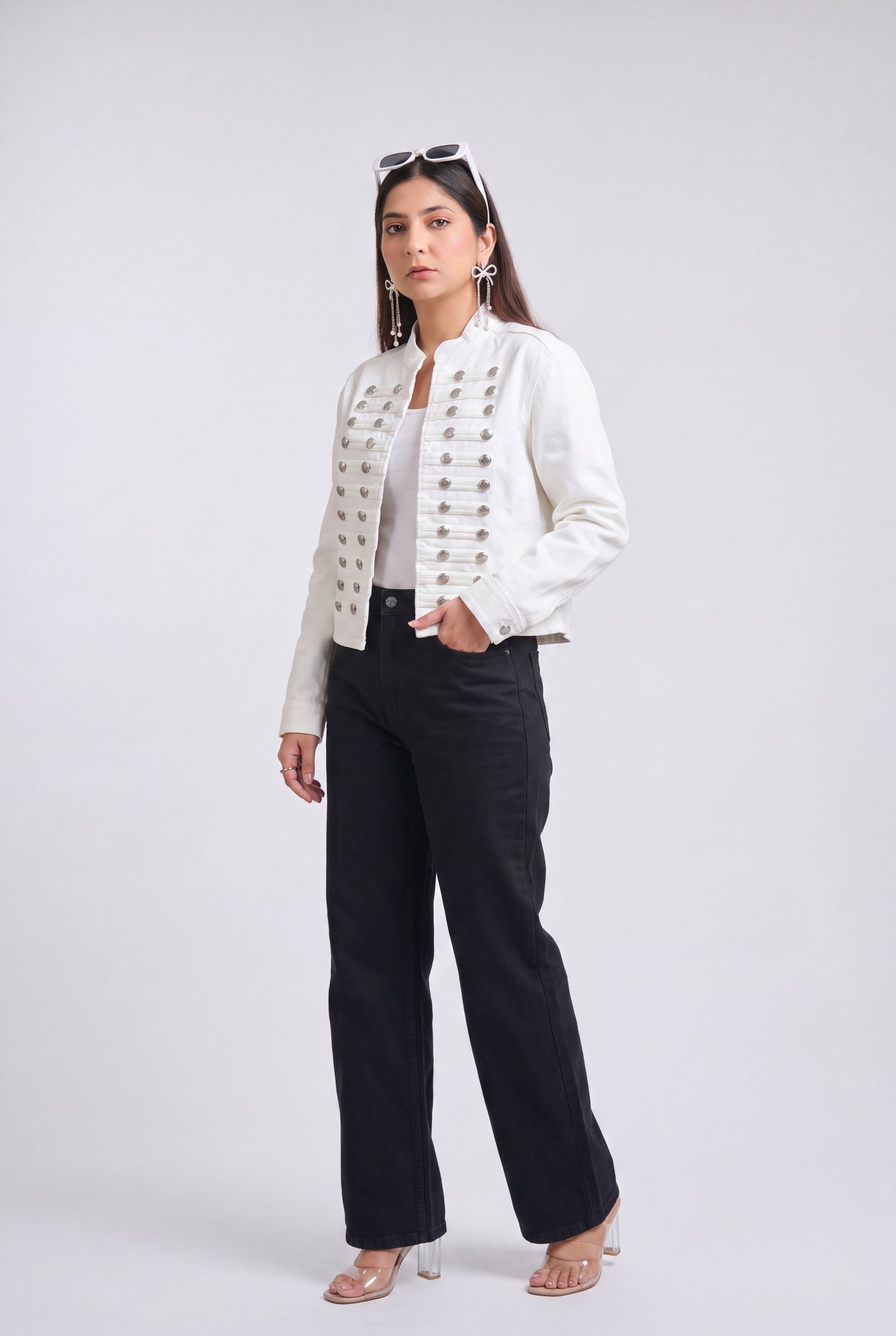 A woman modeling a white cropped jacket with ornate silver button detailing down the front, paired with black wide-leg trousers, against a light gray studio background.
