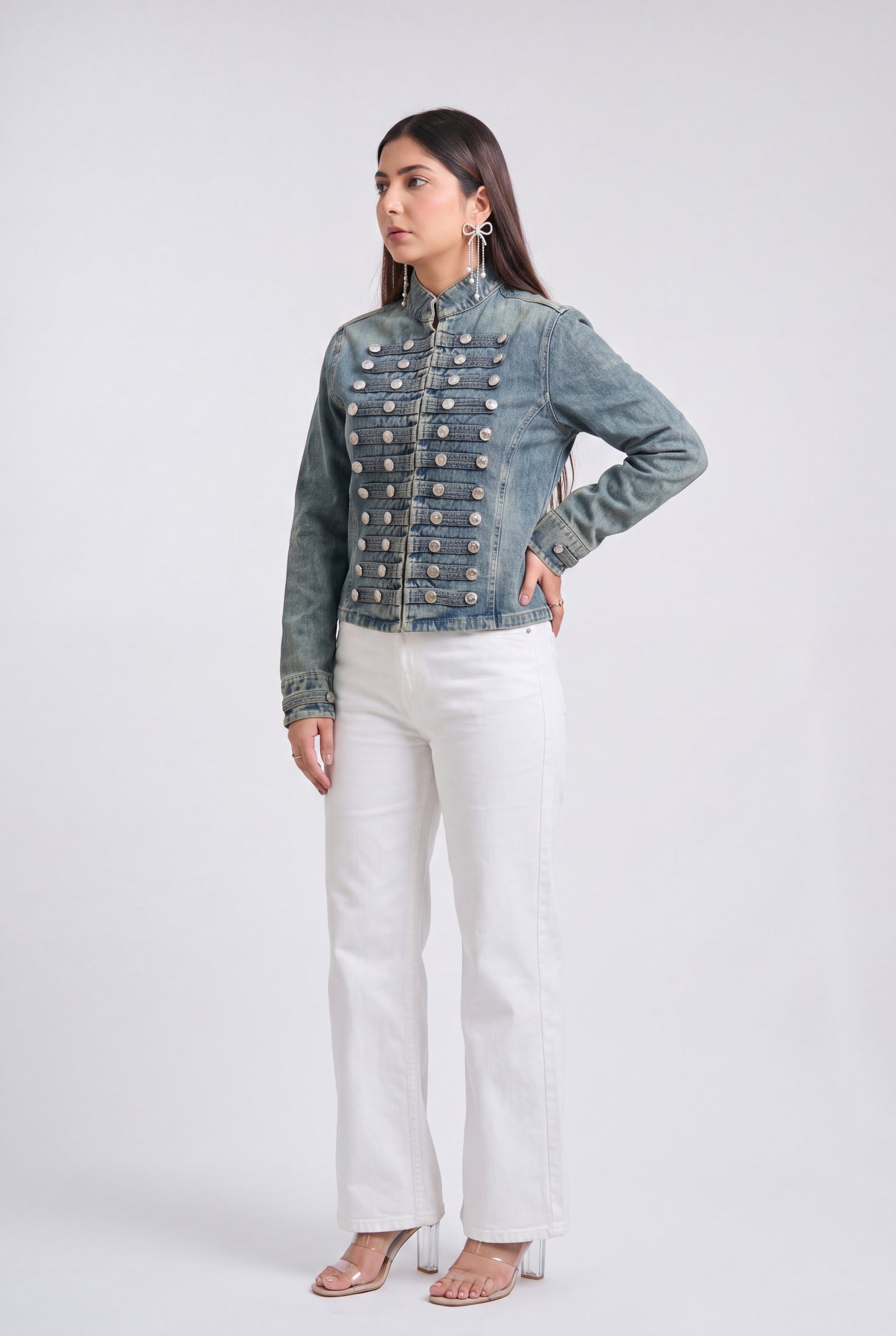 A woman modeling a light blue denim jacket with ornate gold-toned decorative buttons down the front, paired with white pants and clear high-heeled sandals, against a light gray studio background.
