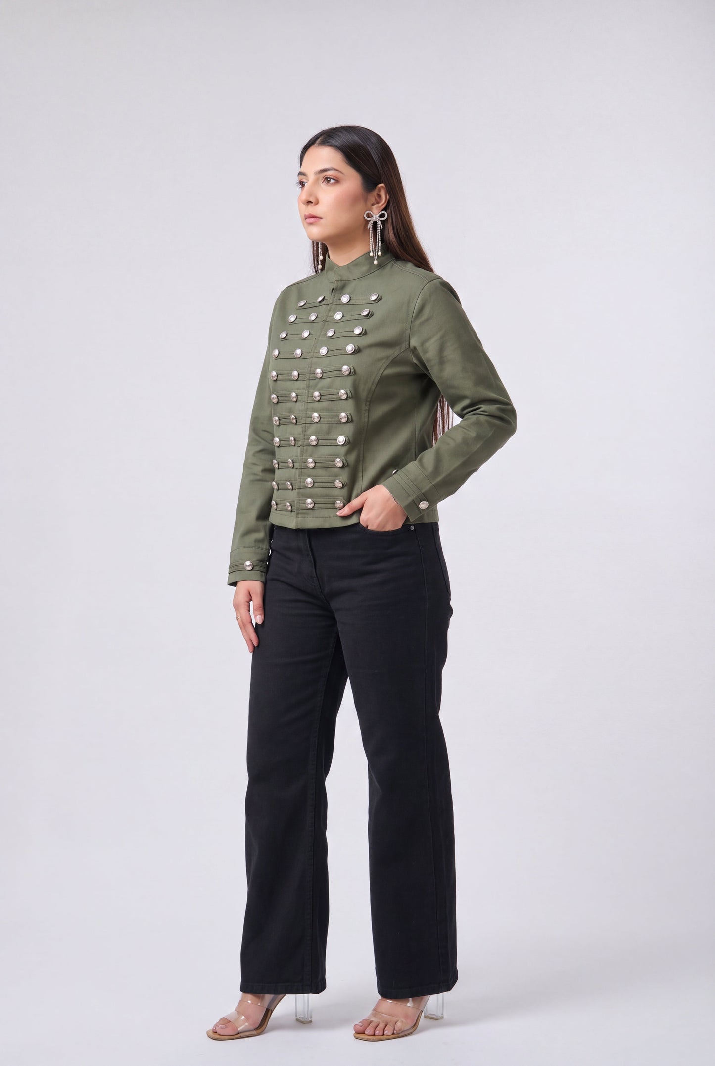 A woman modeling a cropped olive green jacket with silver metallic snap embellishments down the front, paired with black wide-leg pants and clear high-heeled sandals, against a light gray studio background.