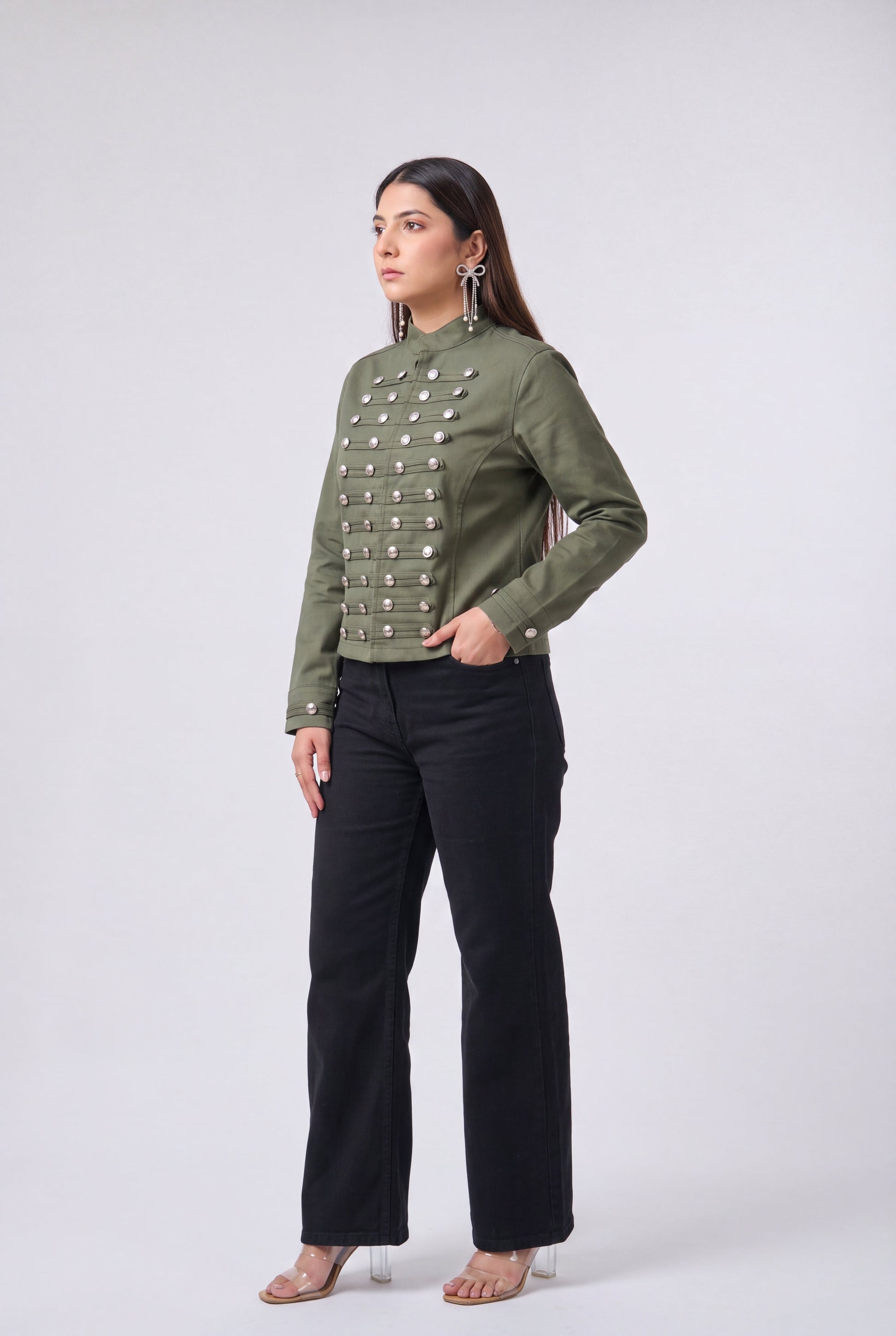 A woman modeling a cropped olive green jacket with silver metallic snap embellishments down the front, paired with black wide-leg pants and clear high-heeled sandals, against a light gray studio background.