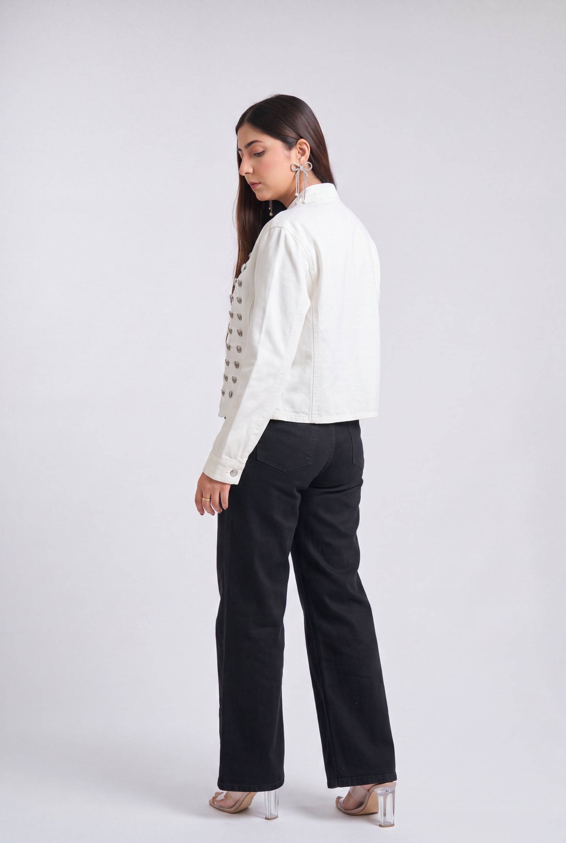 A woman is shown from a side-back angle wearing a white cropped jacket with silver buttons, black wide-leg trousers, and clear high-heeled sandals against a light gray studio background.