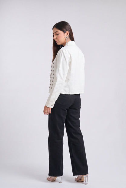 A woman is shown from a side-back angle wearing a white cropped jacket with silver buttons, black wide-leg trousers, and clear high-heeled sandals against a light gray studio background.