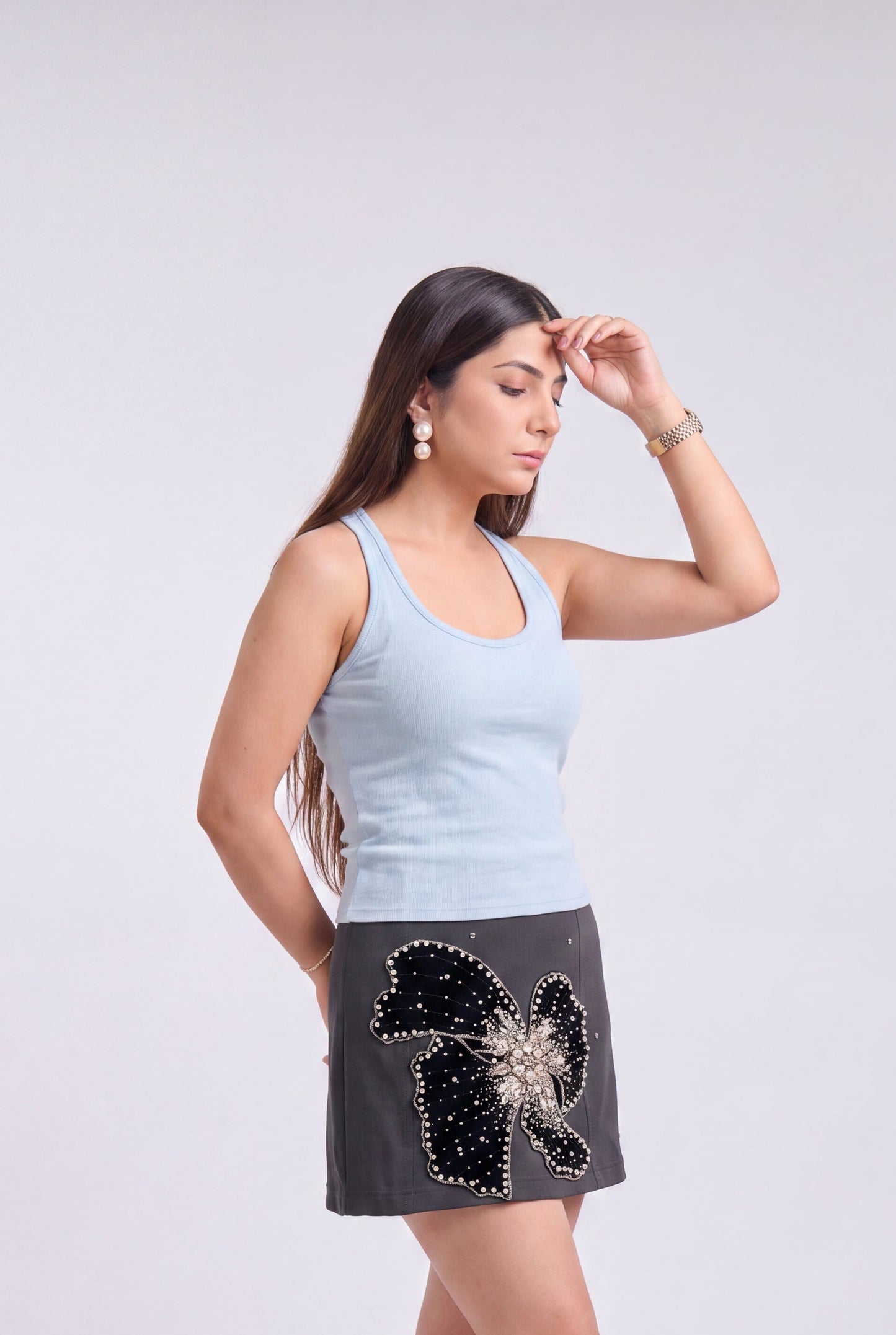 A woman modeling a light blue tank top and a dark gray mini skirt featuring a large black sequined bow and a silver rhinestone flower applique, paired with beige high heels, against a light gray studio background.