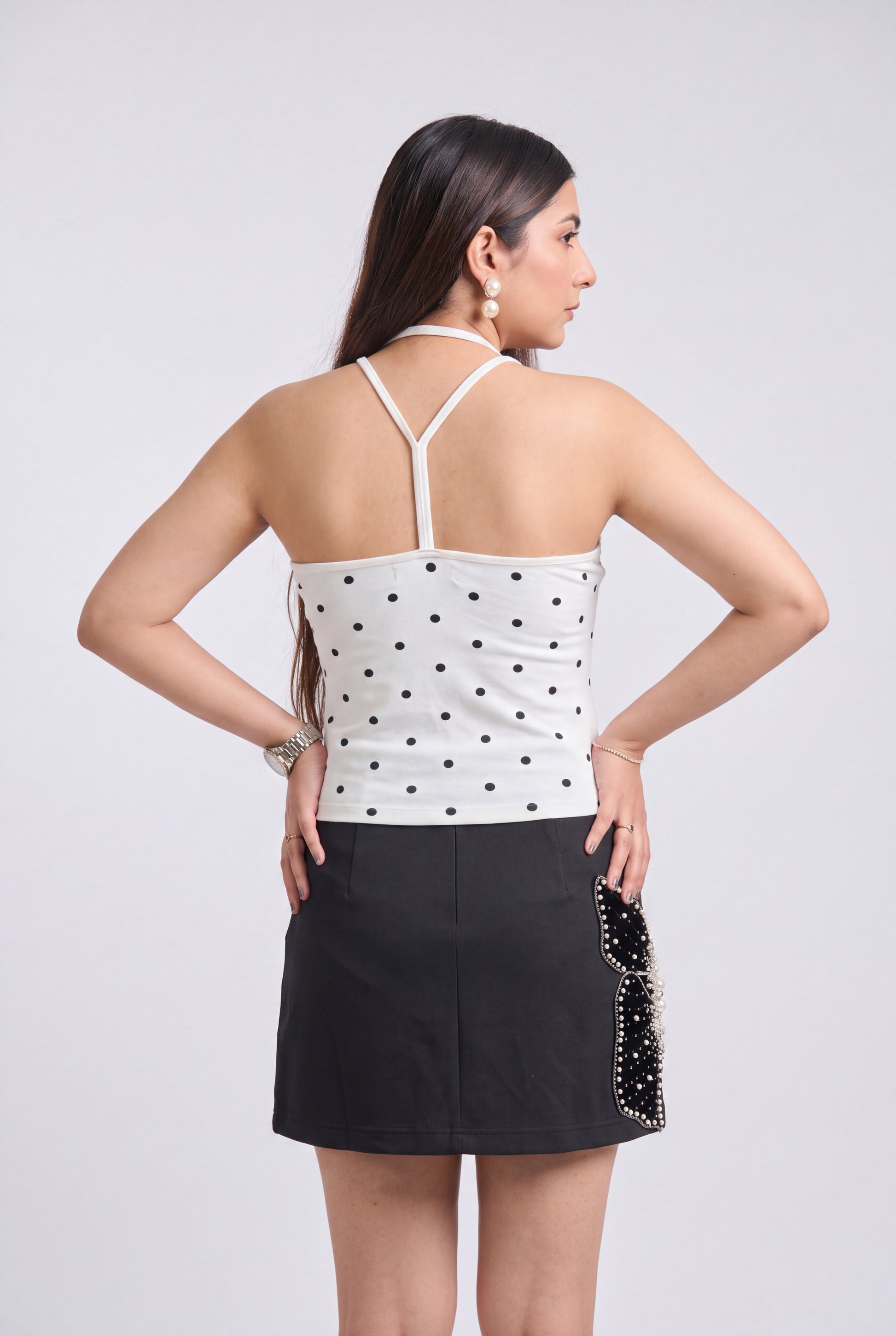 A woman shown from the back wearing a white halter top with black polka dots and a black mini skirt with a black beaded side detail, against a light gray studio background.