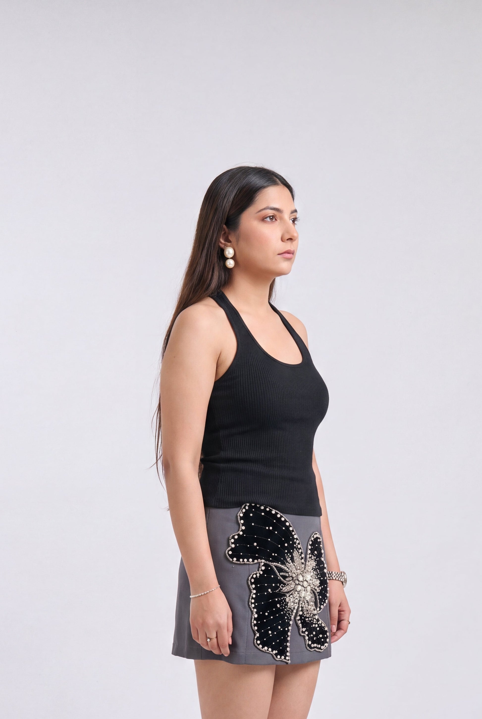 A woman stands in profile wearing a black sleeveless top and a gray mini skirt with a large black and silver sequined butterfly applique on the front, against a light gray studio background.