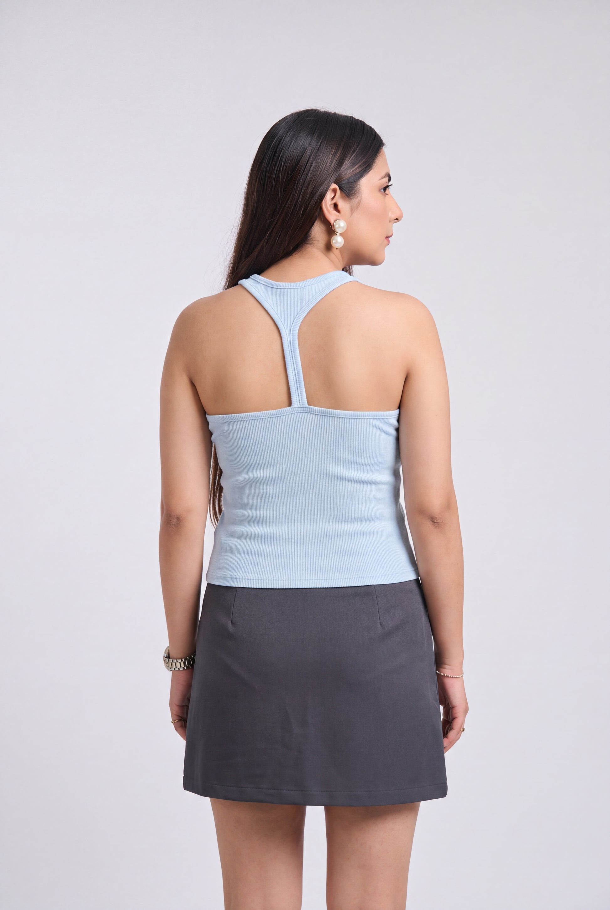 A woman shown from the back wearing a light blue ribbed tank top with a crossover back design and a dark gray mini skirt, set against a plain light gray studio background.