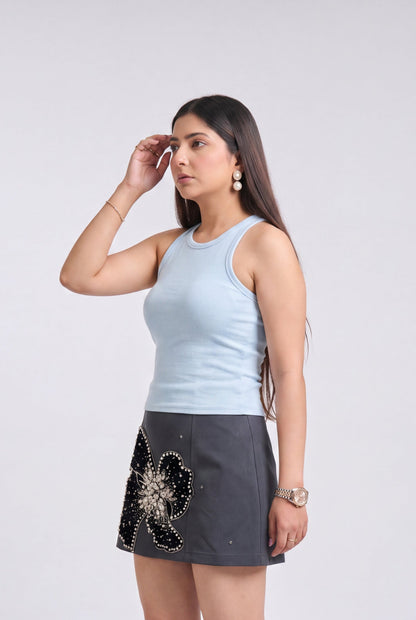 A woman modeling a light blue ribbed tank top and a dark gray mini skirt with a black and silver floral embellishment, paired with beige pointed-toe heels, against a light gray studio background.