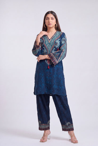A woman modeling a dark blue printed kurta and trouser set with ornate multicolor embroidery on the sleeves and hem, paired with clear high-heeled sandals against a light gray studio background.