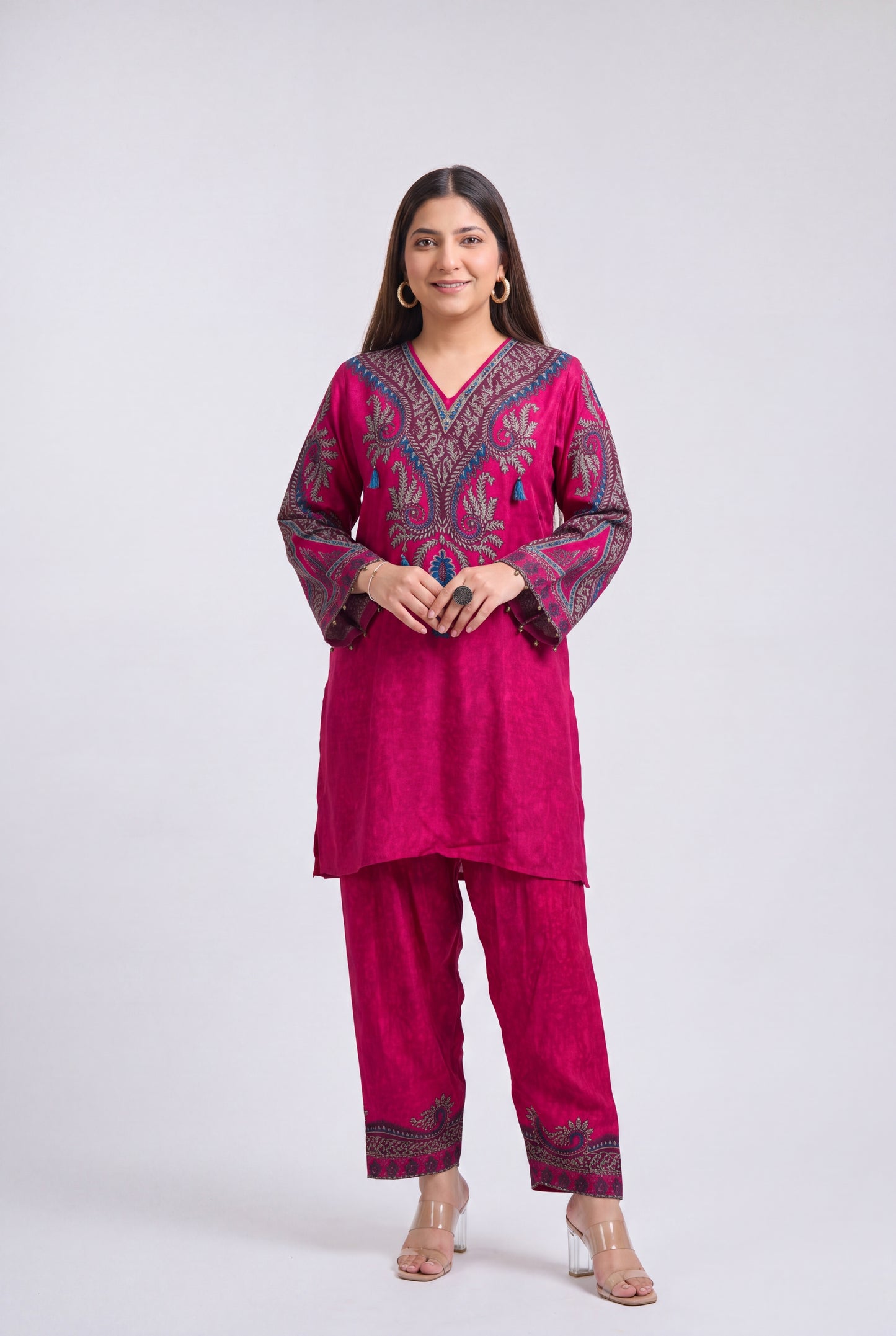 A woman modeling a magenta pink kurta set with dark purple and blue floral embroidery, paired with matching wide-leg trousers, against a light gray studio background.