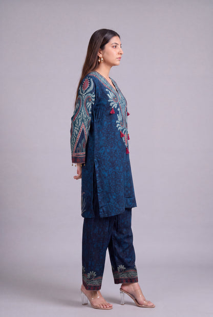 A woman modeling a dark blue printed three-piece traditional outfit with intricate multicolor embroidery on the sleeves and front, paired with matching pants and clear high-heeled sandals, against a solid gray studio background.