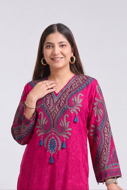 A woman modeling a bright pink traditional kurta set with detailed embroidery, standing next to a wooden stool against a light gray background.