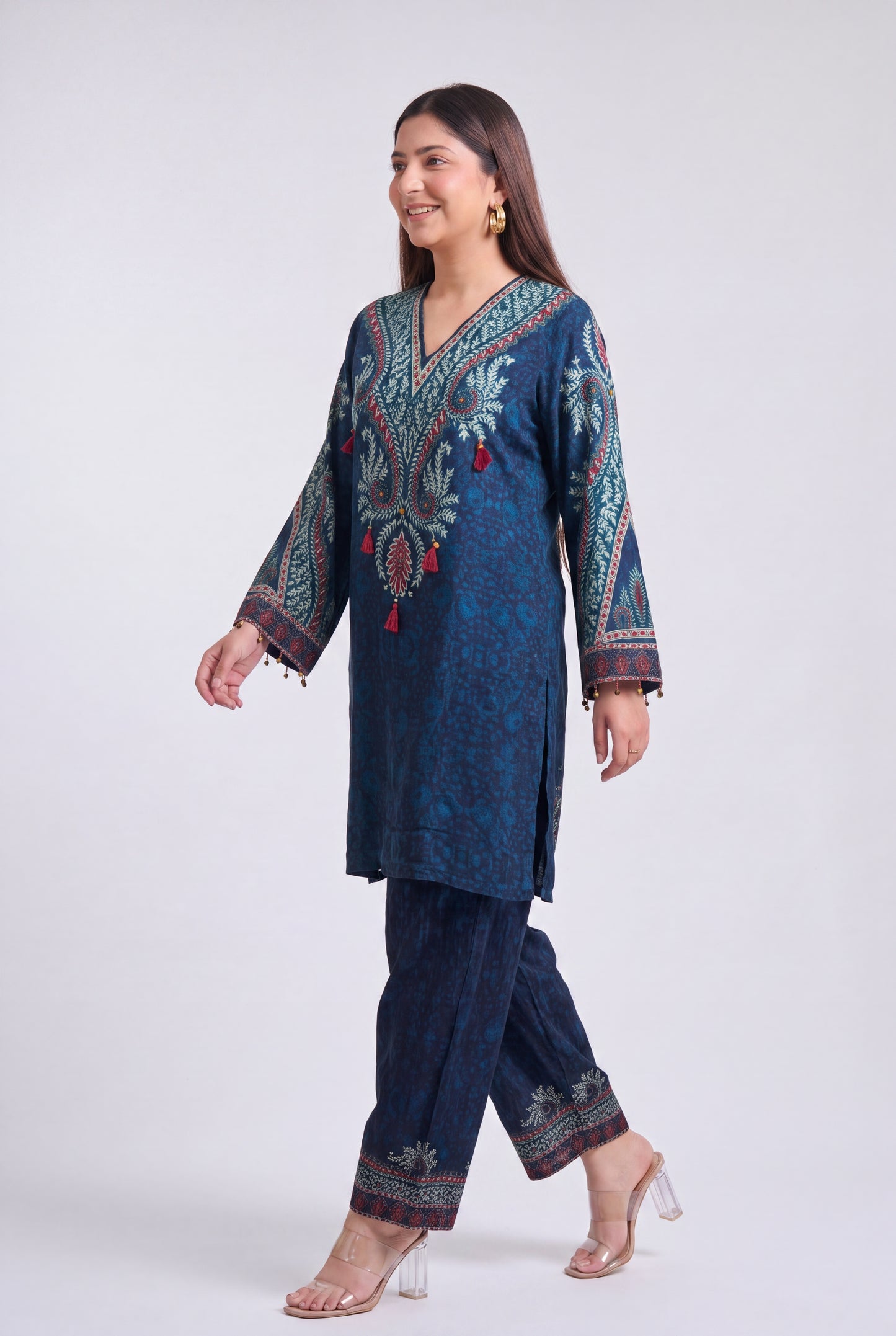 A woman modeling a dark blue printed kurta and matching trousers set with detailed multicolor embroidery and red tassel accents, set against a plain light gray studio background.