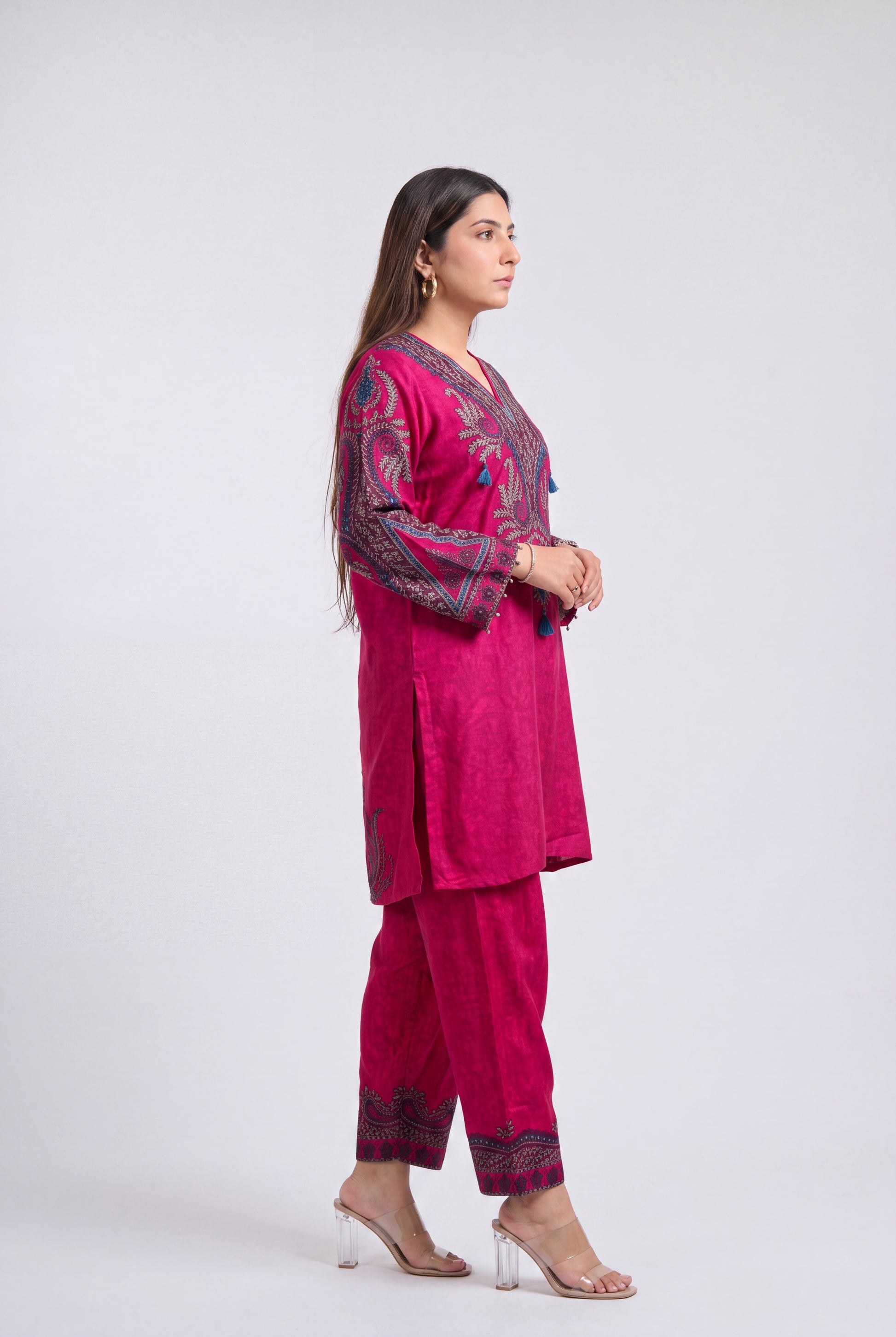 A woman modeling a magenta pink traditional three-piece suit with dark purple and gold embroidery, standing in profile against a light gray studio background.