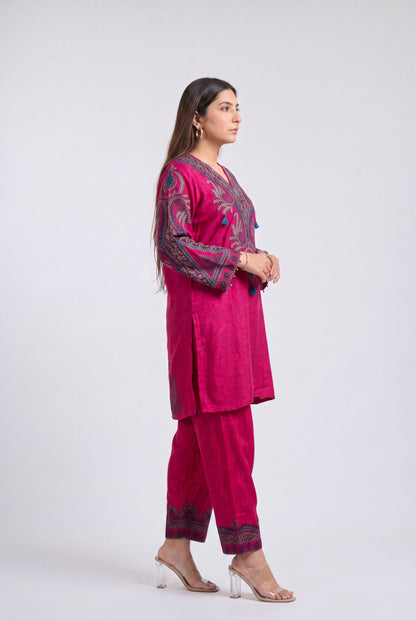 A woman modeling a magenta pink traditional three-piece suit with dark purple and gold embroidery, standing in profile against a light gray studio background.