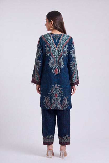 A woman shown from the back wearing a dark blue traditional outfit with intricate multicolor embroidery and patterns, paired with matching pants, against a light gray studio background.
