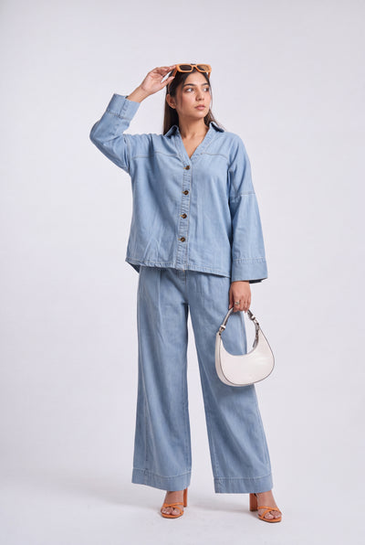 A woman modeling a light blue denim two-piece set consisting of a long-sleeve button-up shirt and matching wide-leg pants, styled with a white crescent handbag and sunglasses against a light gray studio background.