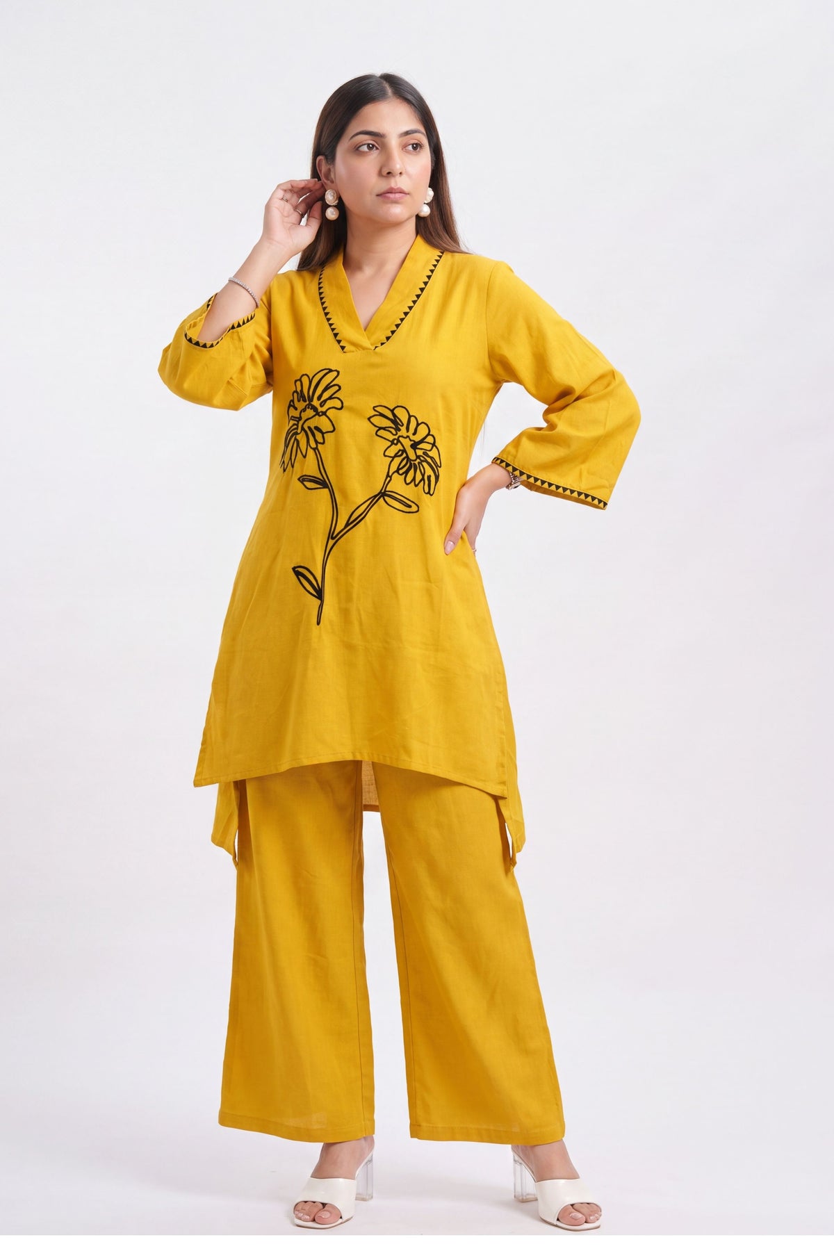 A woman modeling a mustard yellow kurta and trouser set with black floral embroidery on the chest and black trim on the sleeve cuffs, photographed against a plain white studio background.