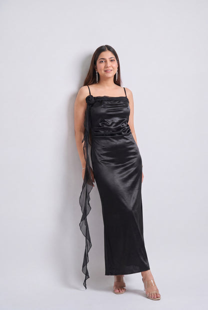 A woman modeling a black satin slip dress with thin straps, a gathered waist, and a long sheer ruffled side drape, standing against a light gray studio background.