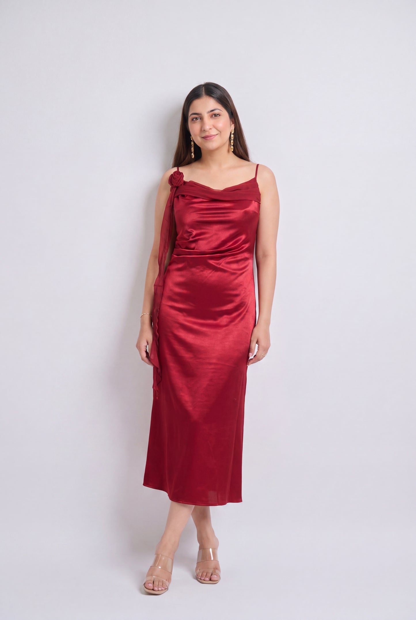 A woman modeling a red satin midi dress with spaghetti straps, a draped neckline, and a side detail, standing against a light gray studio background.