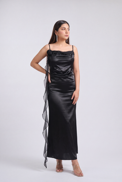A woman modeling a black satin slip dress with thin spaghetti straps, a draped bodice, and a sheer black mesh side drape, photographed against a light gray studio background.