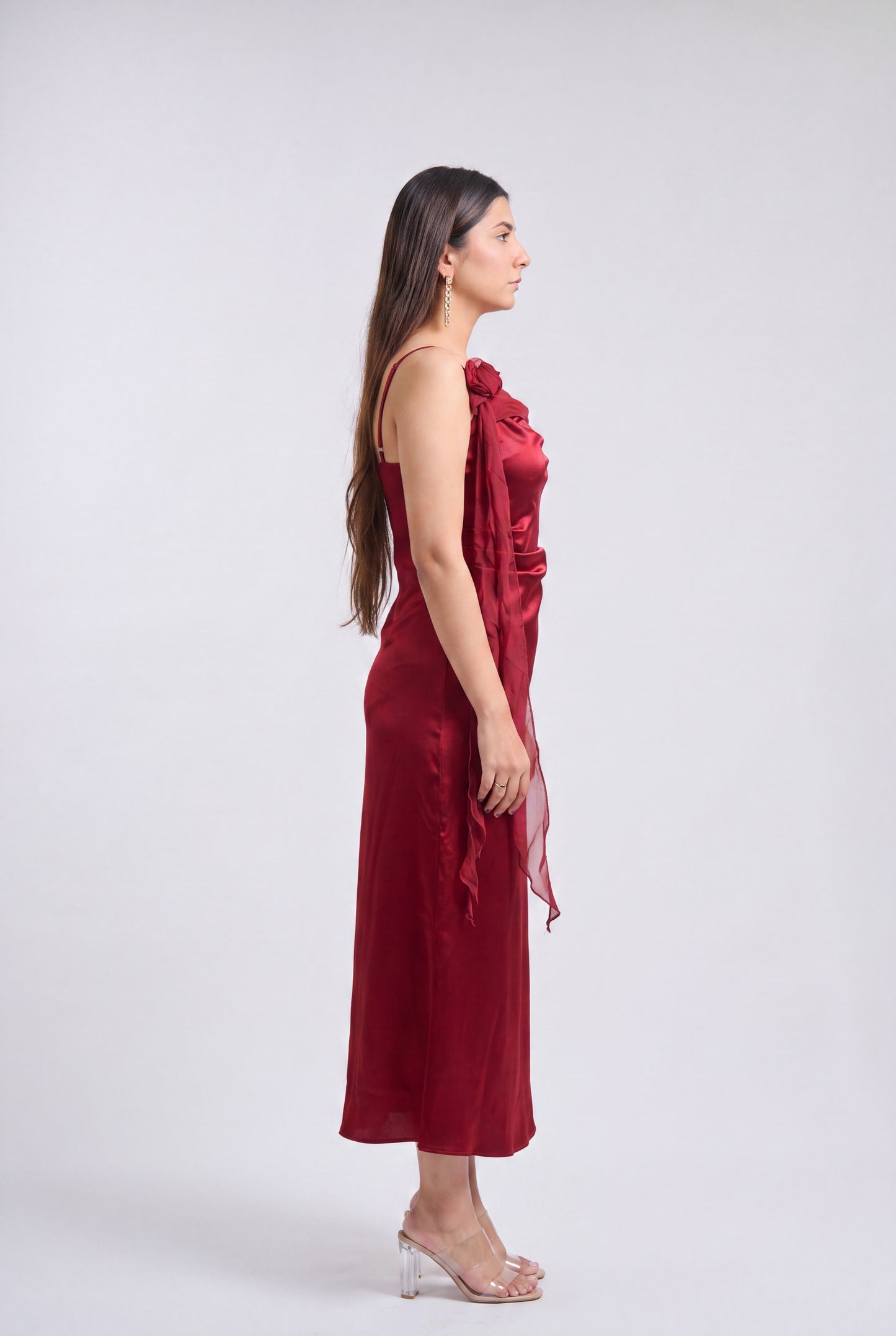 A woman in profile wearing a sleeveless, one-shoulder midi dress in deep burgundy with a draped fabric detail, paired with clear high-heeled sandals, against a light gray studio background.