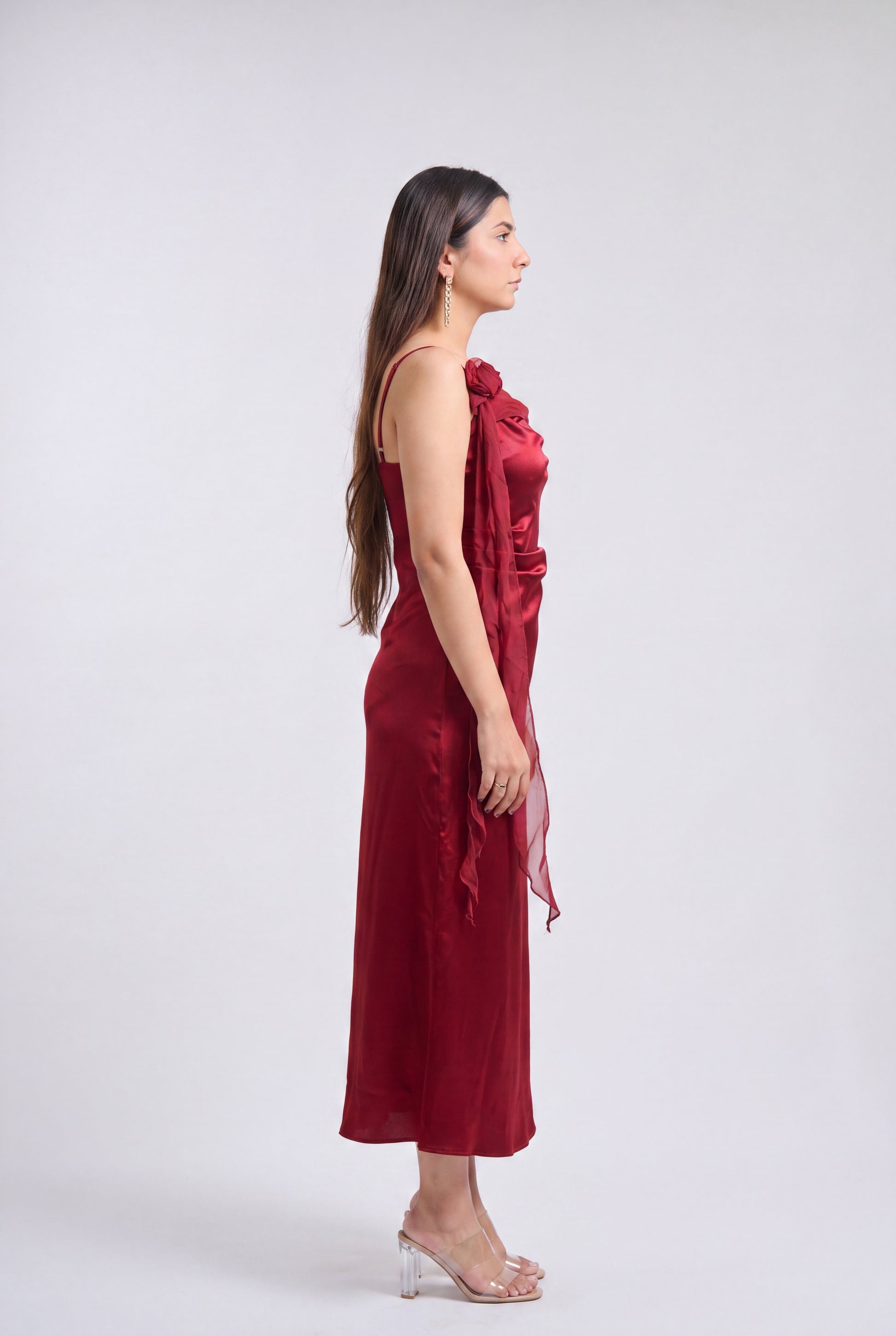 A woman in profile wearing a sleeveless, one-shoulder midi dress in deep burgundy with a draped fabric detail, paired with clear high-heeled sandals, against a light gray studio background.
