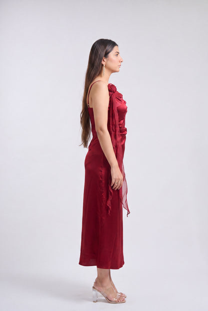 A woman in profile wearing a sleeveless, one-shoulder midi dress in deep burgundy with a draped fabric detail, paired with clear high-heeled sandals, against a light gray studio background.