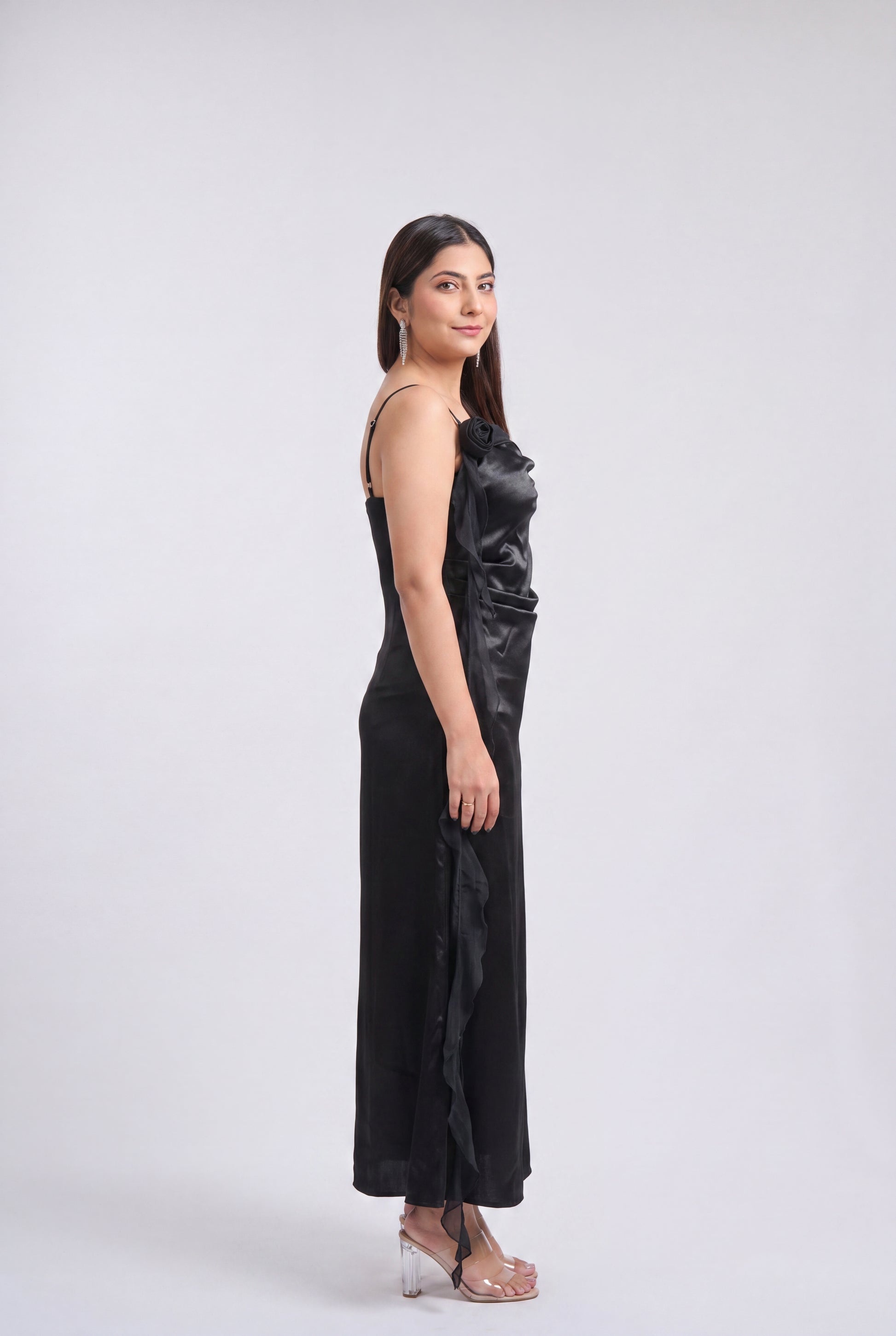 A woman modeling a black satin-like sleeveless maxi dress with a draped, gathered bodice and side slit, standing in clear high-heeled sandals against a light gray studio background.