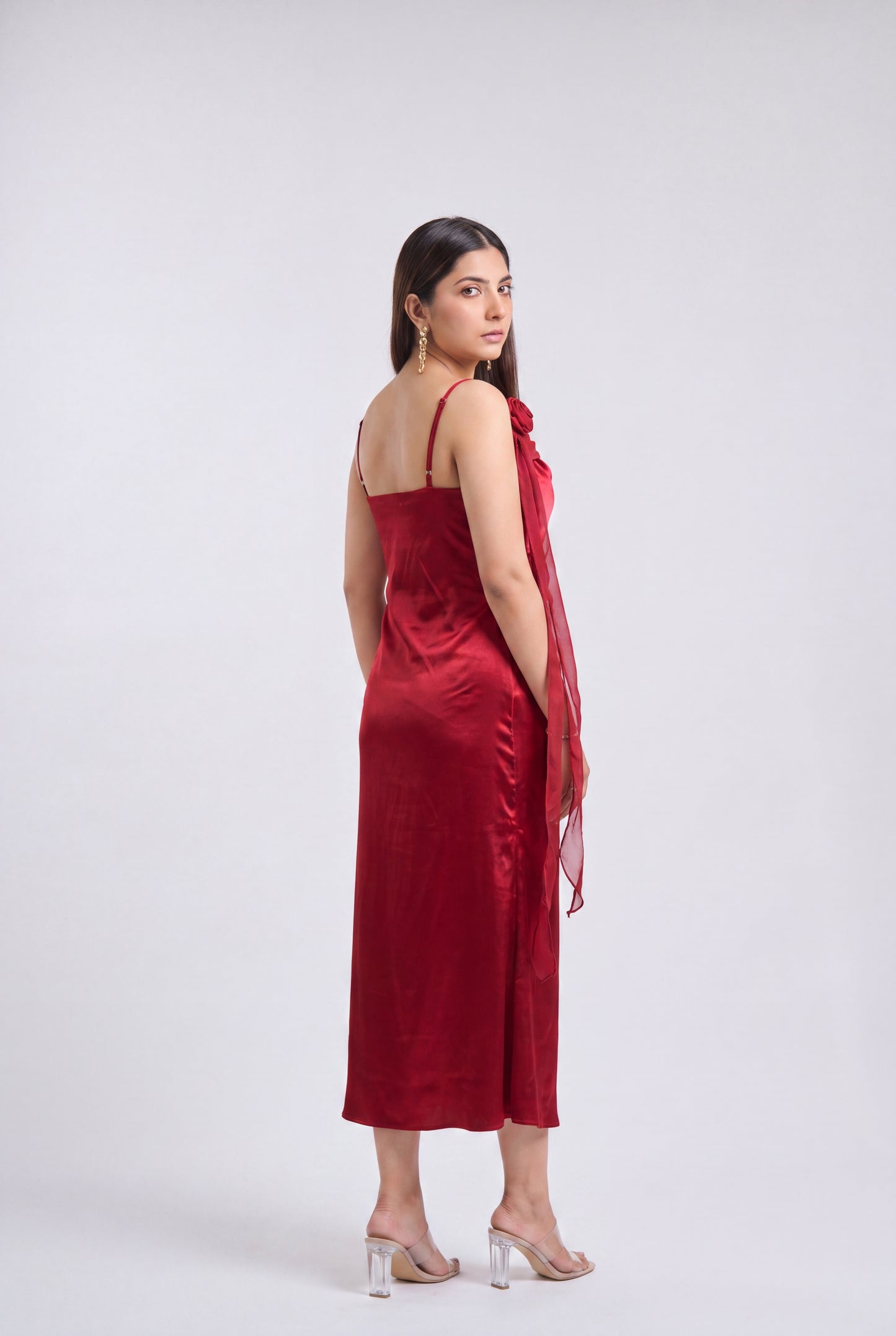 A woman modeling a deep red satin slip dress with thin straps and a draped sheer red shawl, photographed against a neutral light gray studio background.