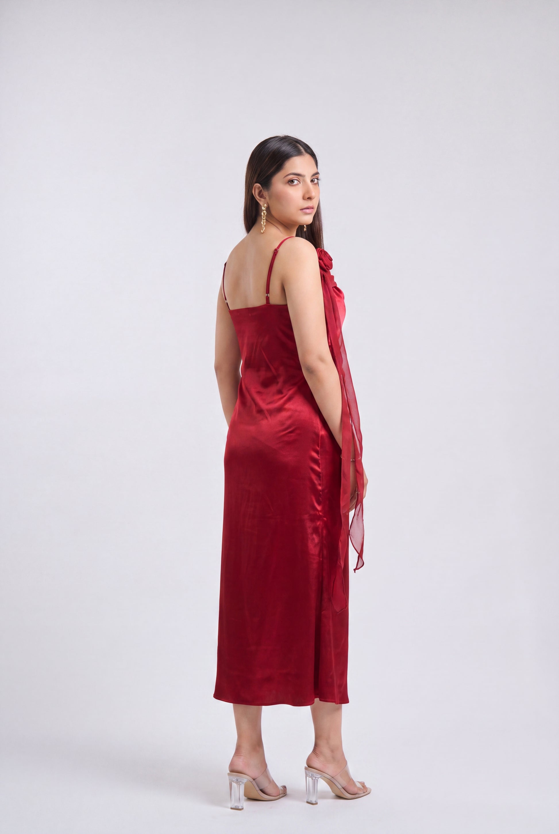 A woman modeling a deep red satin slip dress with thin straps and a draped sheer red shawl, photographed against a neutral light gray studio background.