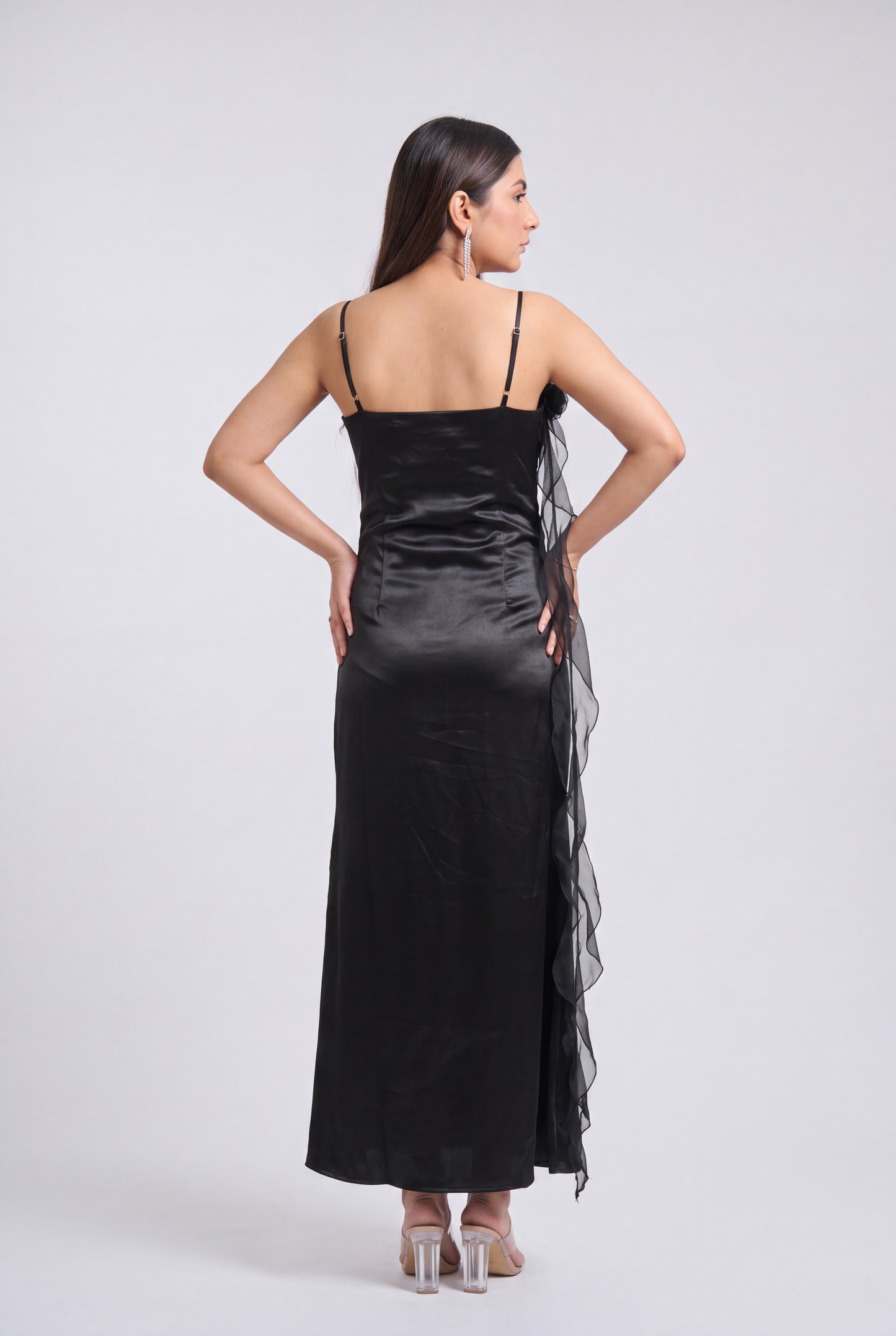 A woman modeling a black satin slip dress with sheer ruffled side panels, shown from the back against a light gray studio background.