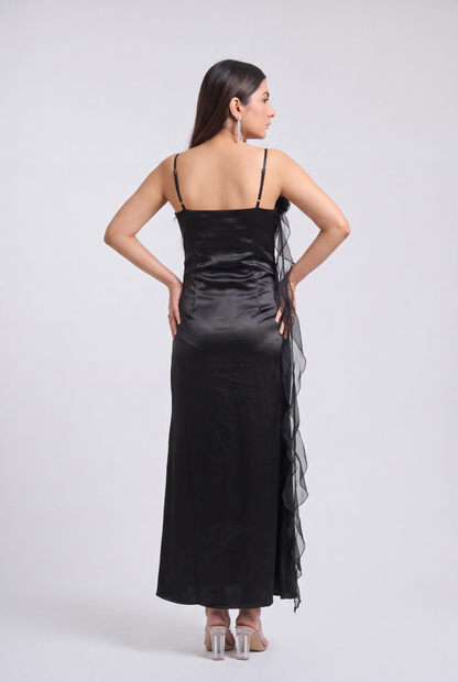 A woman modeling a black satin slip dress with sheer ruffled side panels, shown from the back against a light gray studio background.
