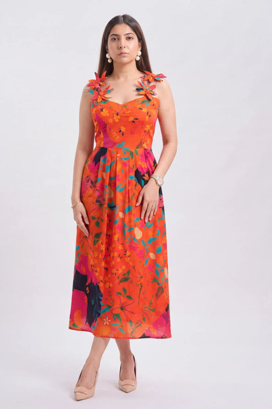 A woman modeling a sleeveless midi dress with a vibrant orange, pink, and teal floral print against a plain light gray studio background.