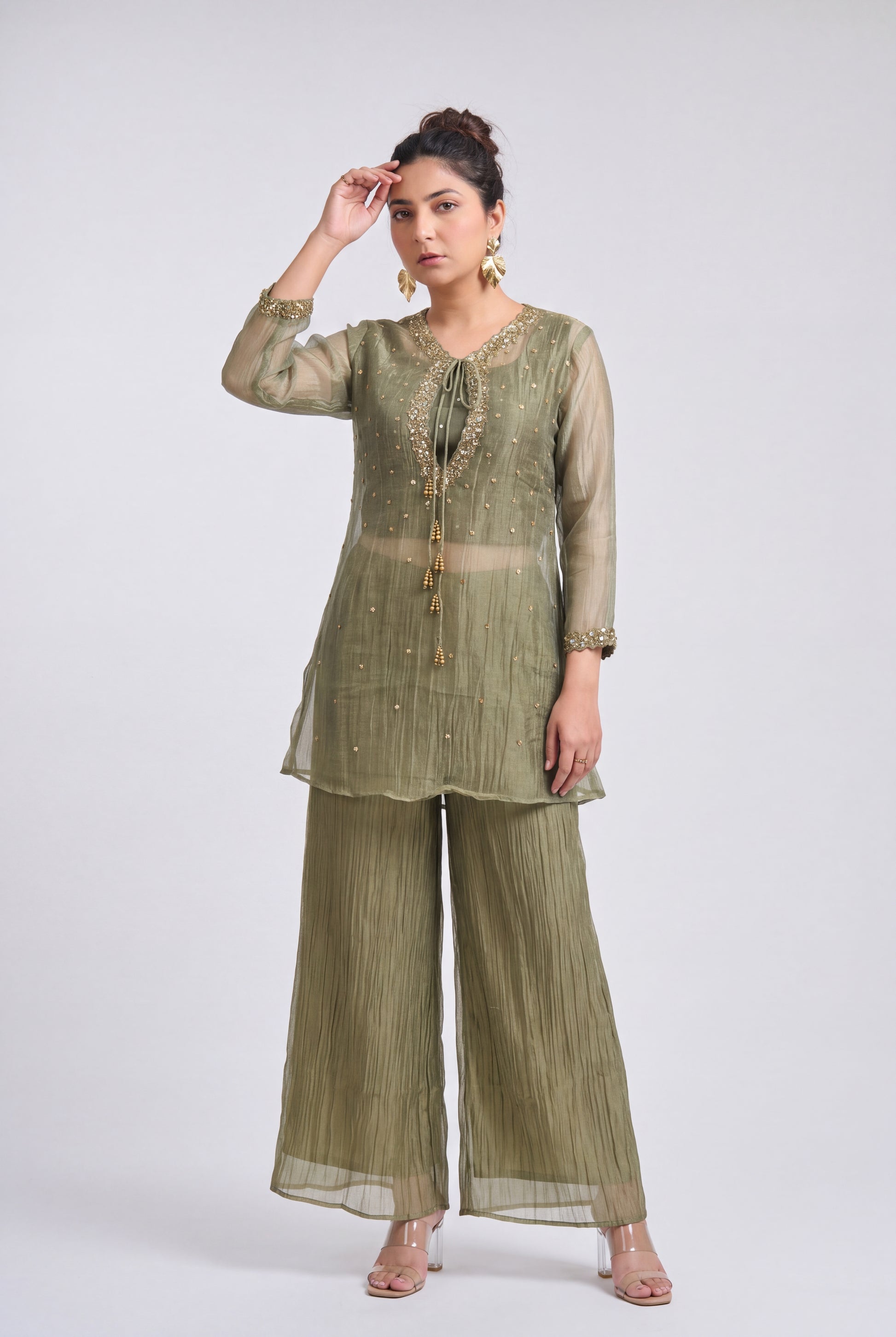A woman models a three-piece olive green ethnic suit consisting of a long-sleeve tunic with gold embroidery, matching pleated wide-leg pants, and sheer matching sleeves, set against a plain light gray studio background.